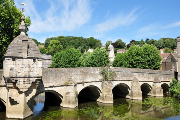 Historic stone bridge over a river with lush greenery and blue sky in the background.