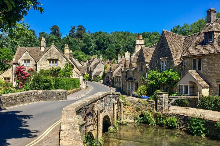 a stone building that has grass in front of a house with Cotswolds in the background