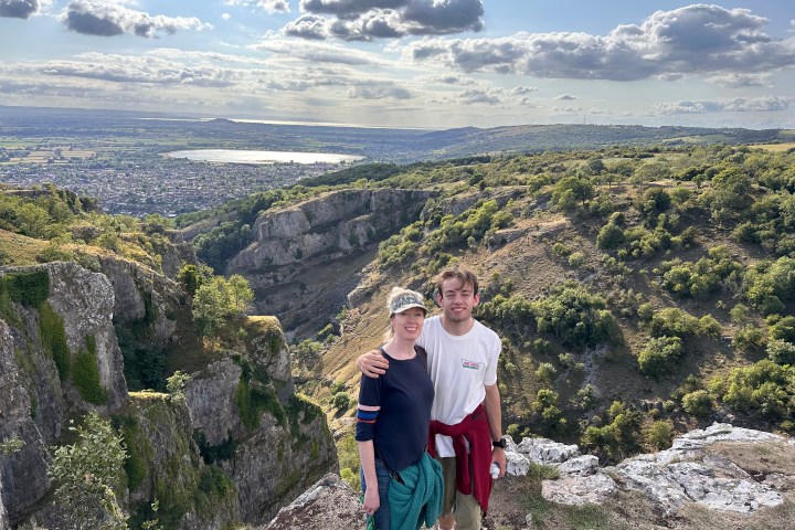 Couple on a cliff with a sweeping valley view under a cloudy sky.