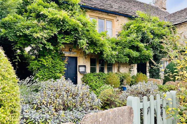 Stone cottage with wisteria-covered facade and a light green picket fence.