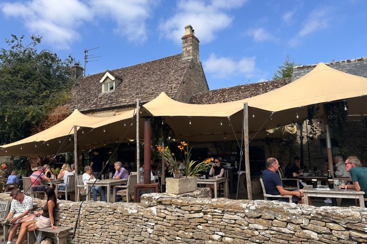 People dining outside under a large beige canopy beside a rustic stone building.