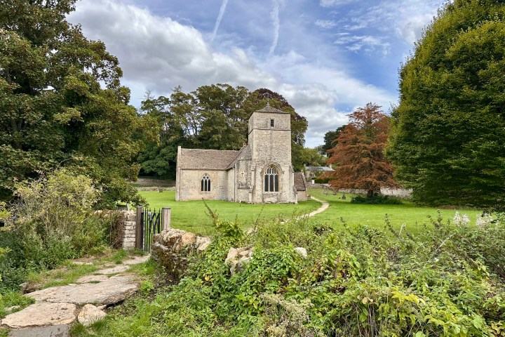 Stone church in grassy field surrounded by trees and a path leading to gate.