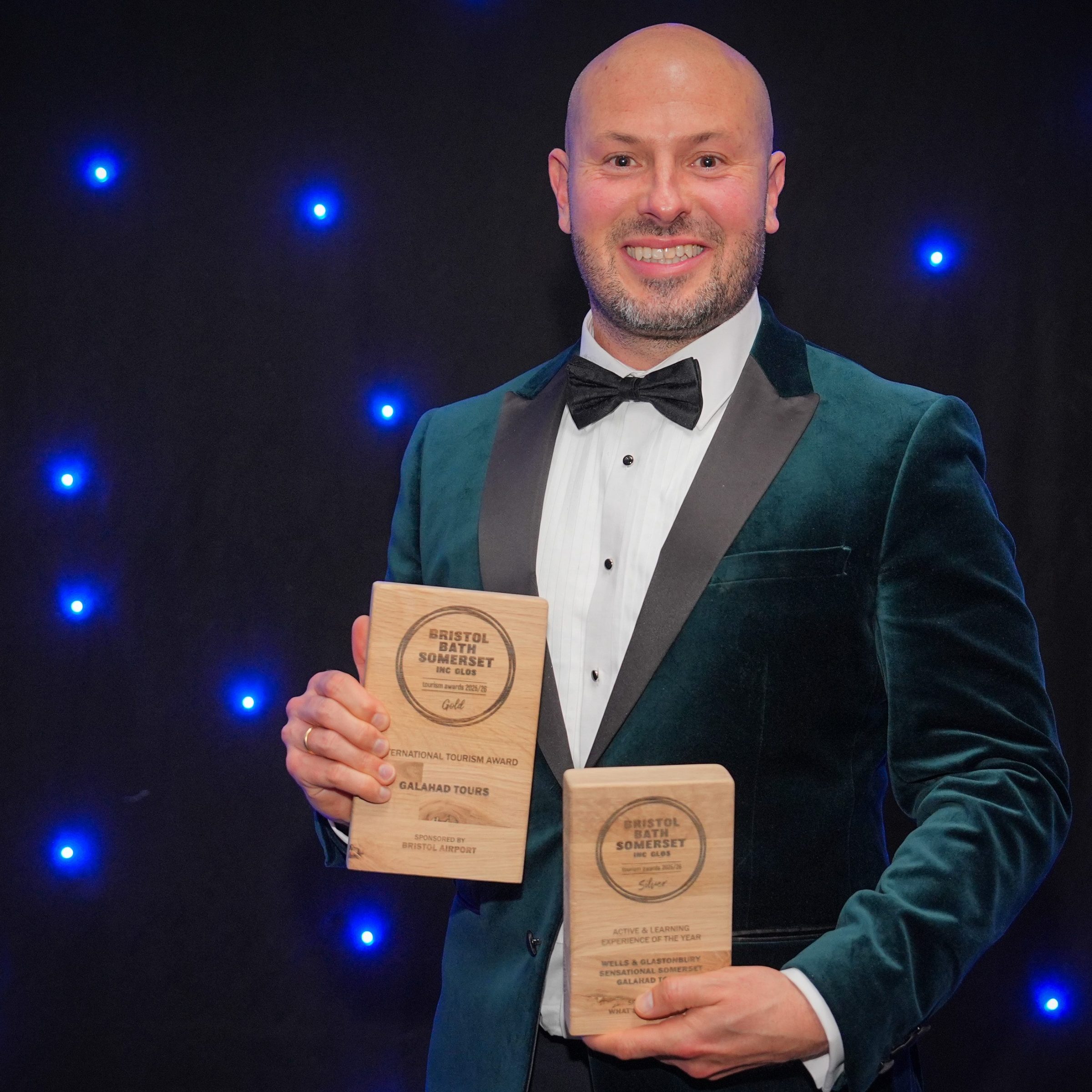 Man in tuxedo holding two awards against a starry blue background.