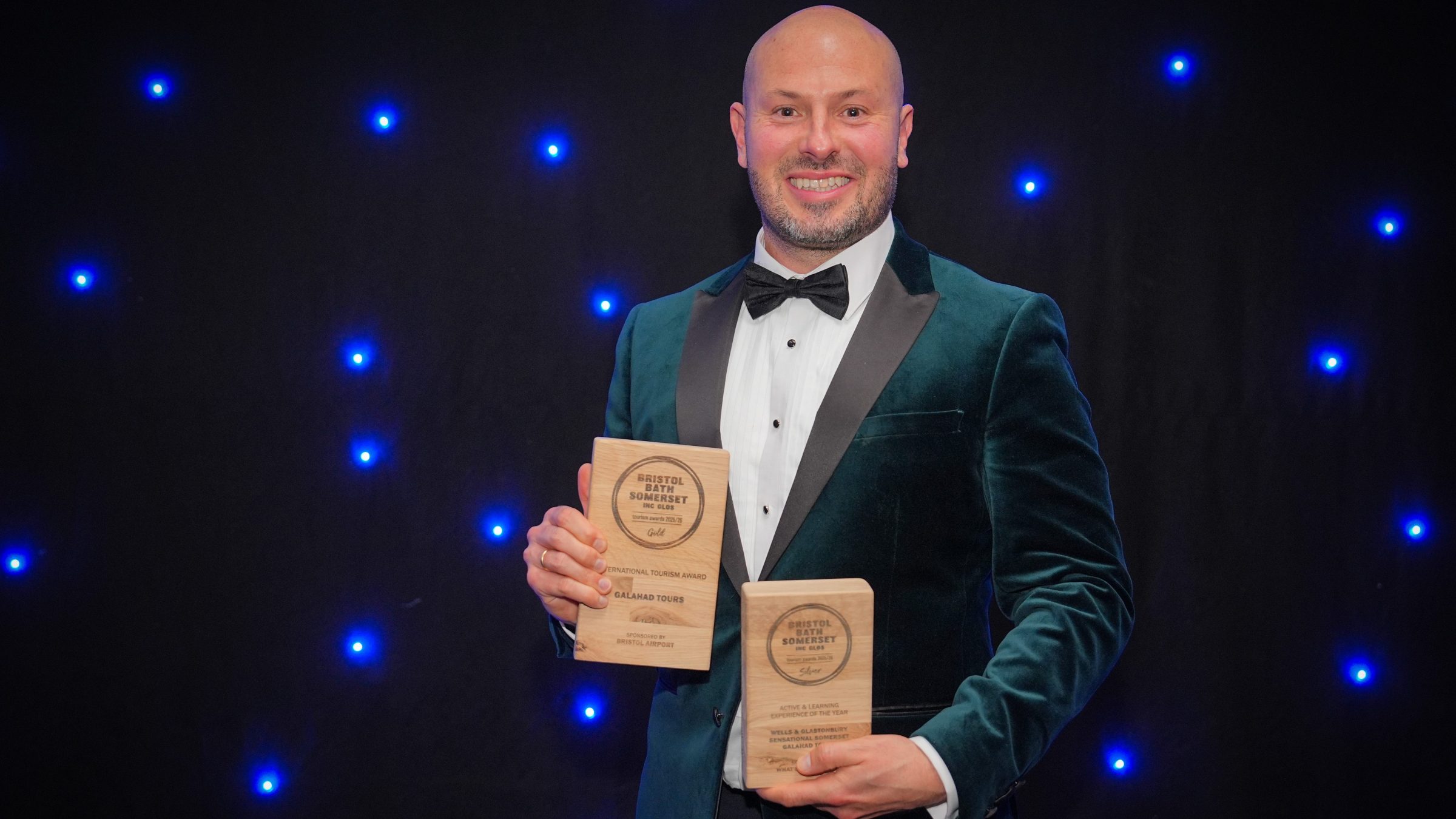 Man in tuxedo holding two awards against a starry blue background.