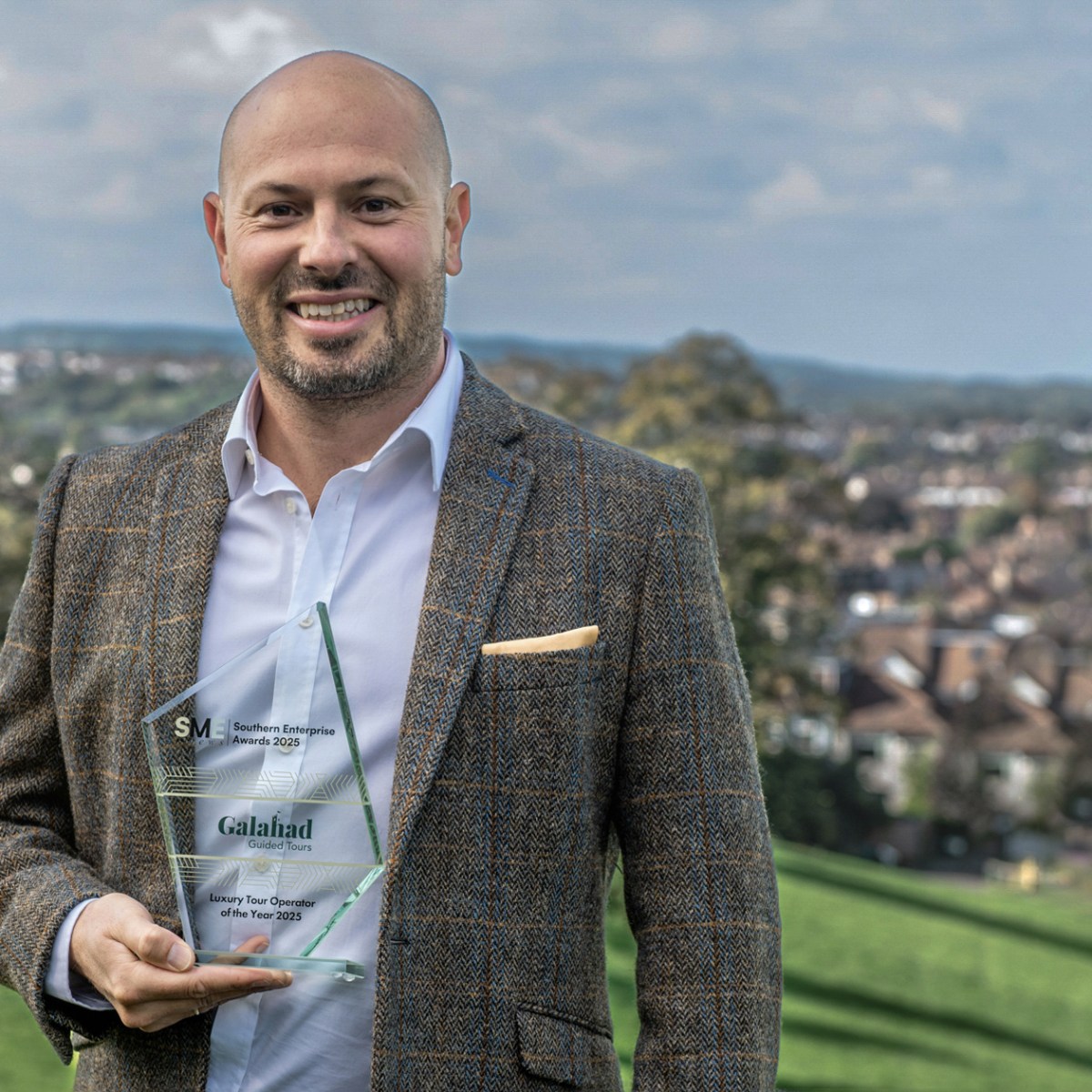 Man holding a trophy, standing on a hill with a town in the background on a cloudy day.