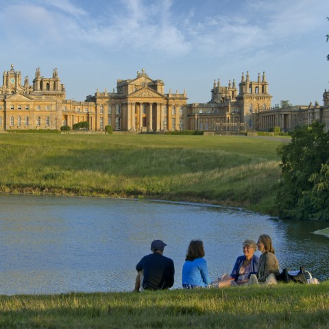 Group of people sitting by a lake with a large historic building in the background.