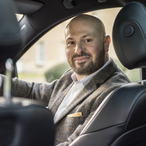Smiling man in a suit jacket sitting in the driver's seat of a car, looking towards the camera.