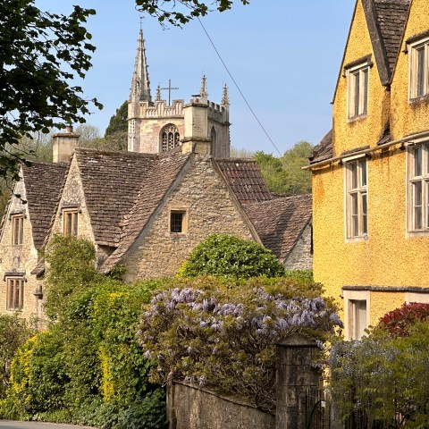 Charming village street with stone houses, flowering bushes, and a church steeple in the background.