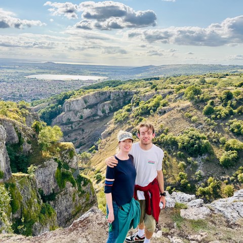 Two people posing on a rocky hilltop with a scenic valley and sky in the background.