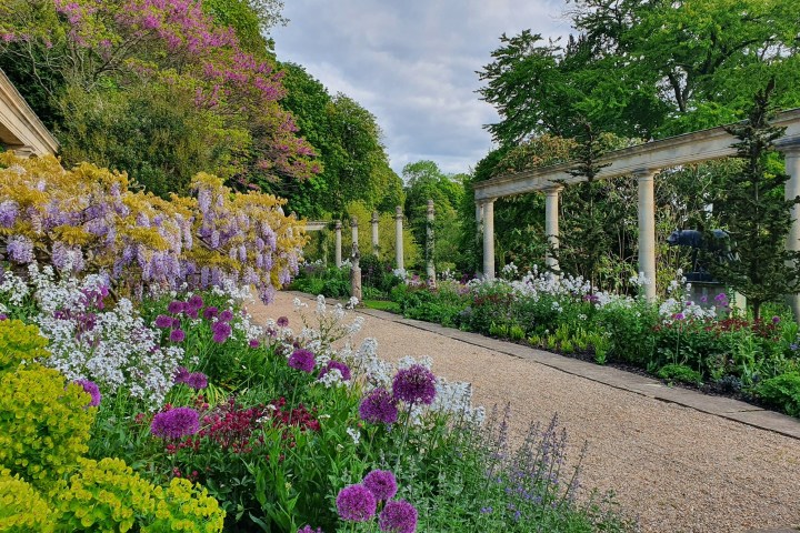 Colorful garden with flowers, trees, and stone columns along a gravel path.