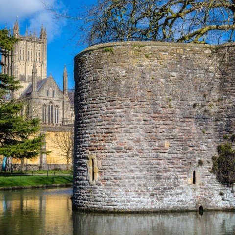 Ancient stone wall with a moat, cathedral tower and trees in the background on a sunny day.