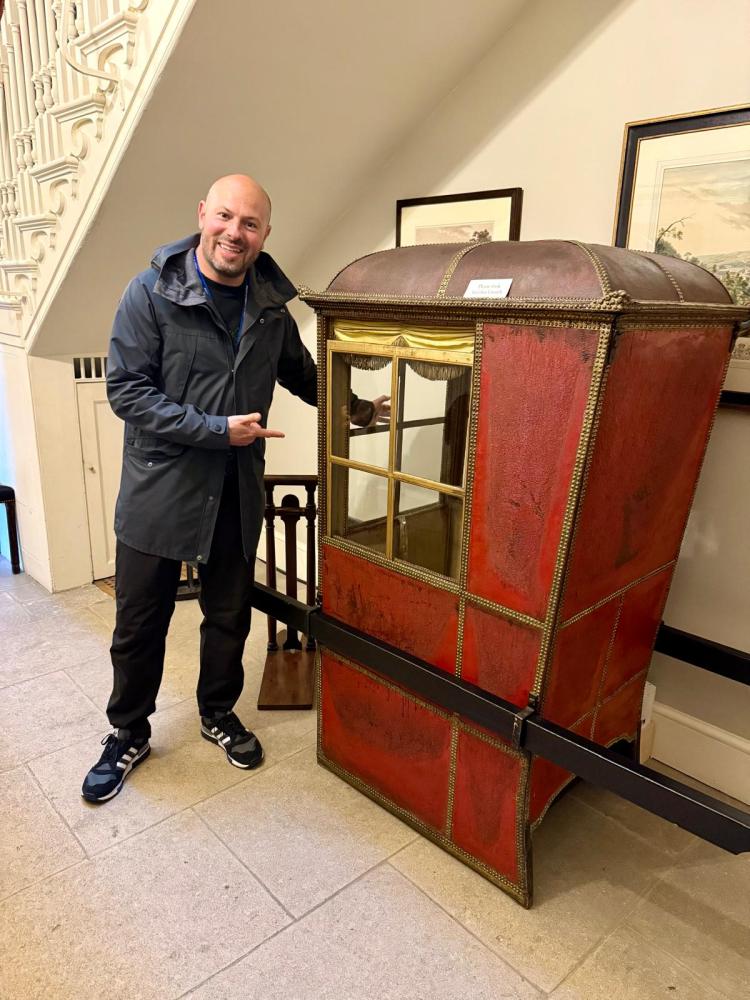 Person smiling and pointing at a vintage red sedan chair indoors.