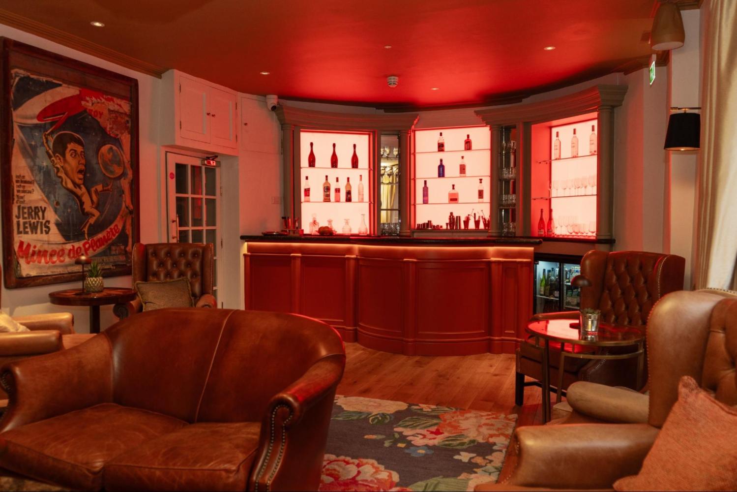 Cozy bar with leather chairs, red-lit shelves with bottles, and a vintage poster on the wall.