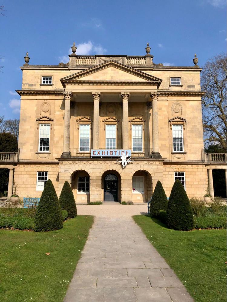 Historic building with columns, exhibition banner on facade, pathway and hedges leading to entrance.