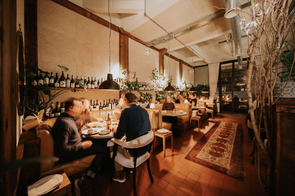 Cozy restaurant interior with people dining, warm lighting, and shelves of wine bottles.